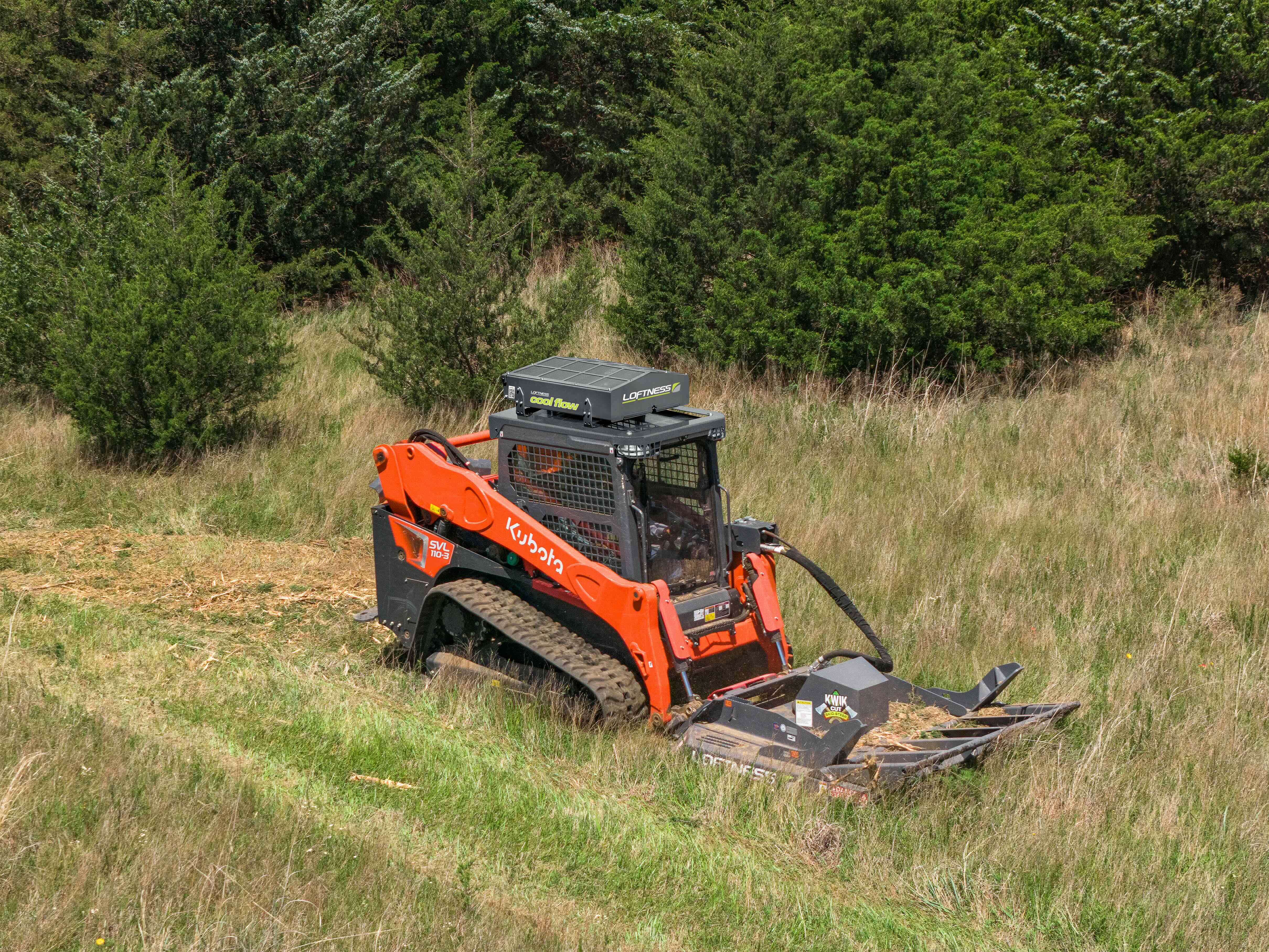 Orange Kubota skid steer loader clearing overgrown field with brush attachment near forest