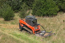 Orange Kubota skid steer loader clearing overgrown field with brush attachment near forest