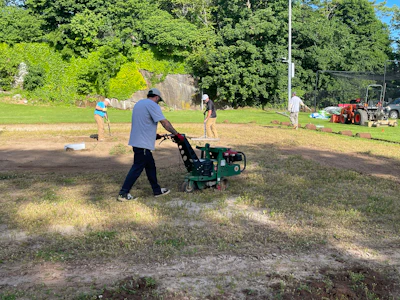 Person operating a green walk-behind aerator machine on a grassy field during outdoor maintenance work