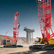 Heavy construction cranes and crawler carriers on dusty job site with bridge pillar under clear sky