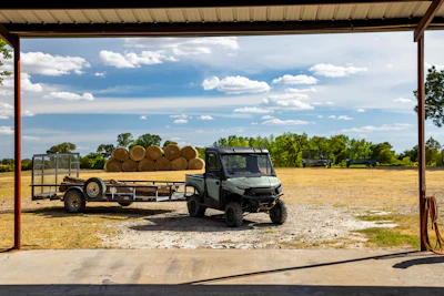 Black utility vehicle with hay-loaded trailer parked under metal structure in rural farmland with trees and blue sky