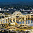 Aerial view of a yellow arch bridge spanning a blue river with city skyline and forested landscape in background