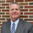 Professional headshot of a man in gray suit and striped tie against brick background