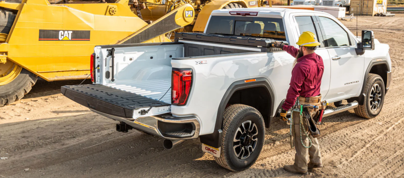 Construction worker in yellow hard hat standing beside white GMC pickup truck with open bed on job site with yellow CAT bulldozer