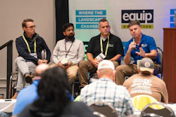 Four panelists seated on stage during a conference discussion with audience members in foreground and branded backdrop signage behind them.