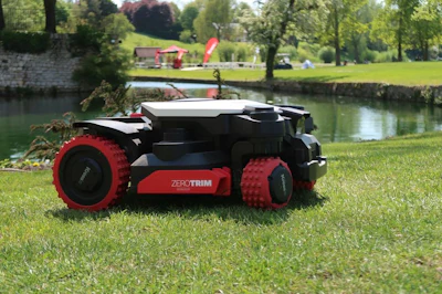 Red and black robotic lawn mower with ZEROTRIM branding on grass by a river