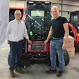 Two men standing in front of red Takeuchi construction equipment in industrial warehouse