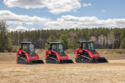 Three red tracked skid-steer loaders lined up in a sandy field with pine forest in background