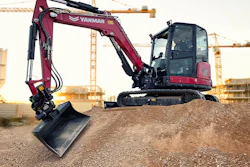 Burgundy YANMAR mini excavator with extended arm on gravel pile at construction site with tower cranes