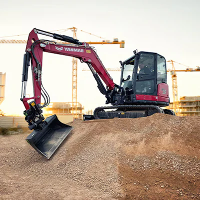 Burgundy YANMAR mini excavator with extended arm on gravel pile at construction site with tower cranes