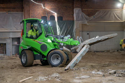 Bright green compact wheel loader with operator moving wooden beam on construction site