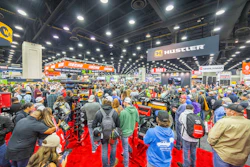 Crowded trade show floor with hundreds of attendees browsing equipment and product booths under bright overhead lighting