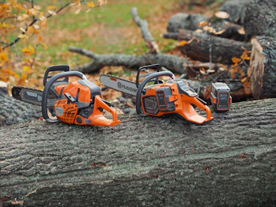 Two orange and black Husqvarna chainsaws positioned on a weathered log with forest debris in background