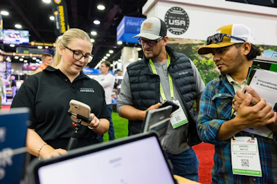 Three professionals at a trade show booth discussing equipment, with laptops and branded signage visible