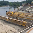 Waterfront construction site with yellow cranes, concrete structures, and workers near a river surrounded by trees
