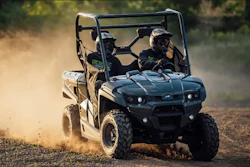 Two riders operating a black and tan UTV on a dusty off-road trail with protective gear and helmets
