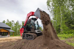 Red tracked skid steer loader dumping soil at a construction site near a building and forest