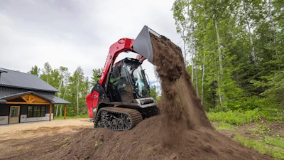Red tracked skid steer loader dumping soil at a construction site near a building and forest
