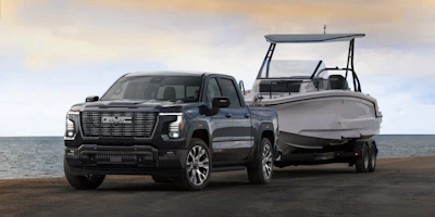Black 2027 GMC Sierra 1500 Denali Ultimate pickup truck towing a white boat on a beach with ocean backdrop
