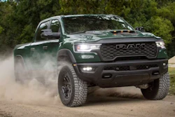 Dark green RAM truck kicking up dust on a dirt road with trees in background