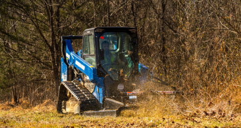 Blue tracked skid steer loader clearing brush in autumn forest