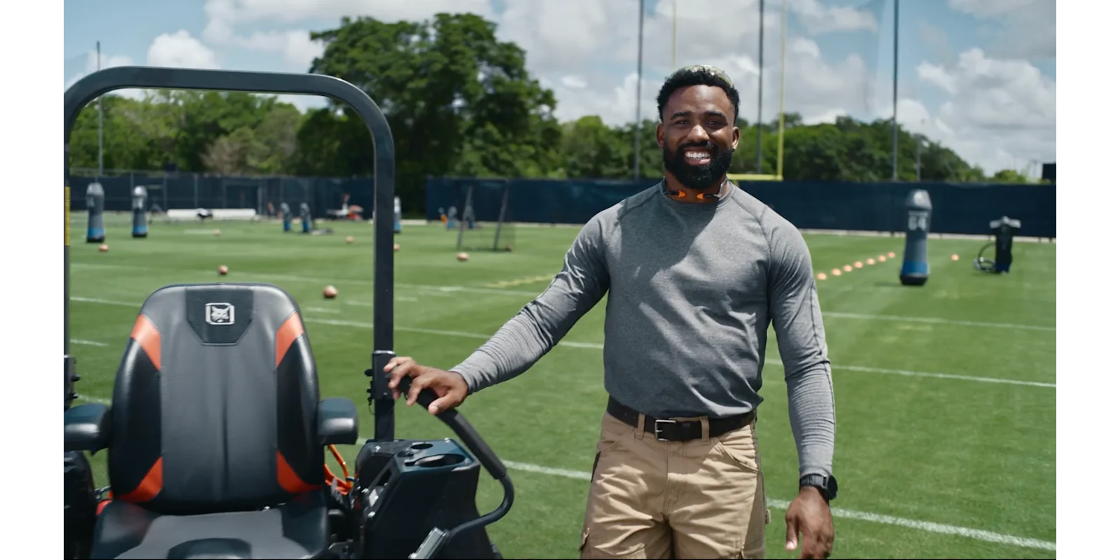 Man smiling while standing with a riding mower on a sports training field with goal posts and cones
