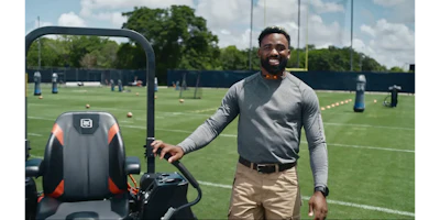 Man smiling while standing with a riding mower on a sports training field with goal posts and cones