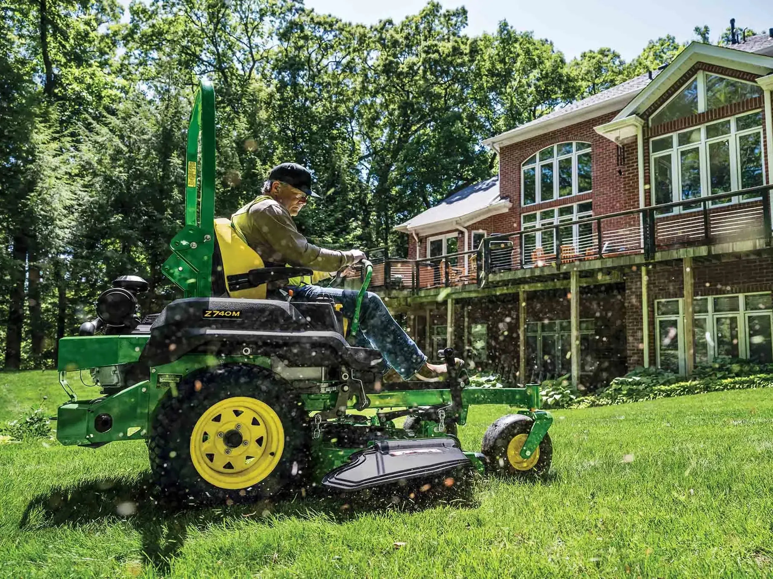 Man operating green and yellow John Deere lawn mower on residential property with brick house and trees