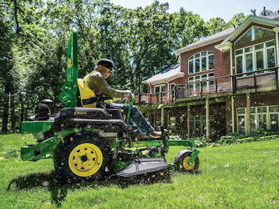 Man operating green and yellow John Deere lawn mower on residential property with brick house and trees