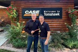 Two men in dark shirts stand in front of wooden building with CASE Construction and Welcome to Tomahawk signage