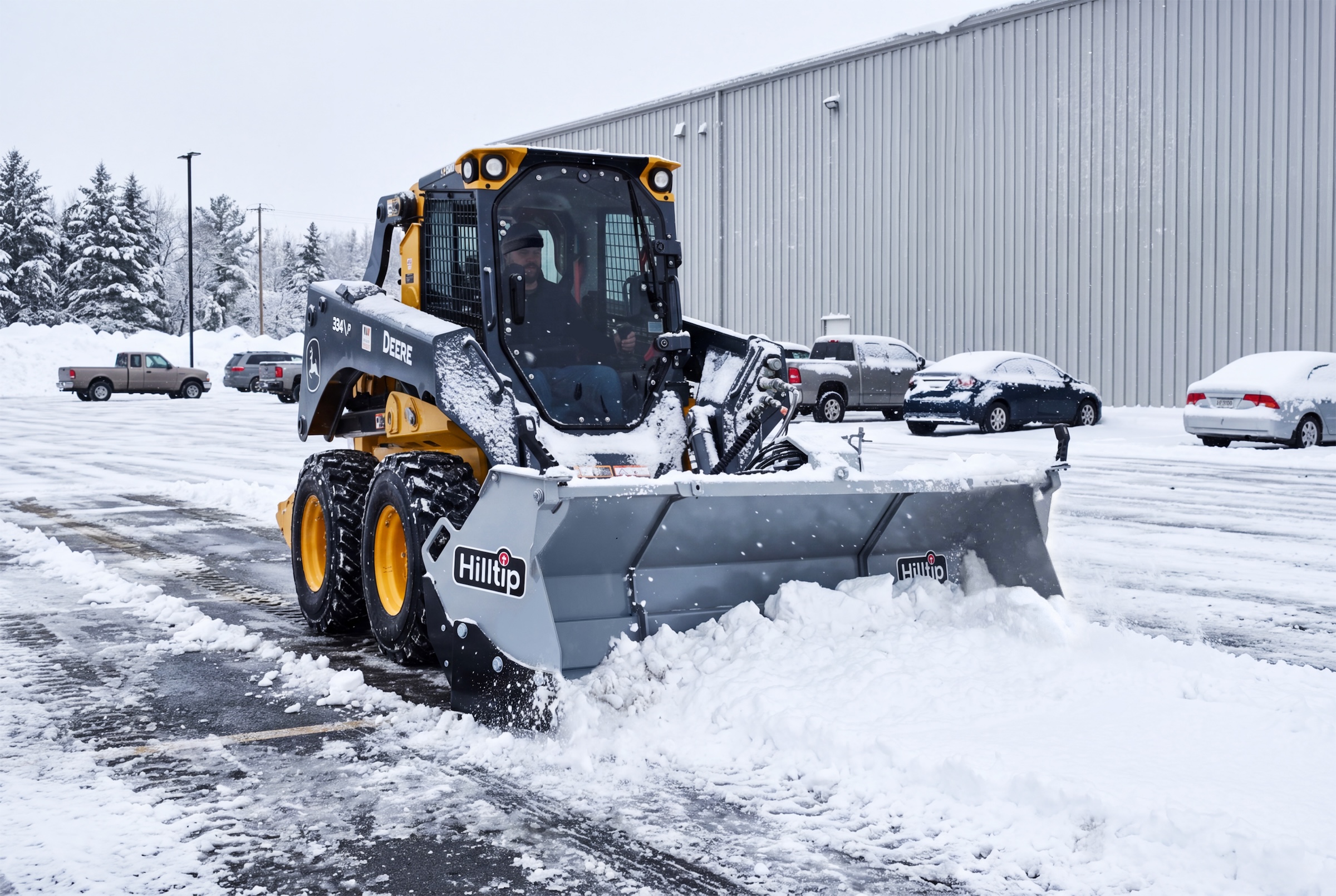 Yellow and black snow plow clearing parking lot during winter with vehicles and industrial building in background