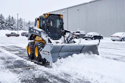 Yellow and black snow plow clearing parking lot during winter with vehicles and industrial building in background