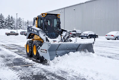Yellow and black snow plow clearing parking lot during winter with vehicles and industrial building in background