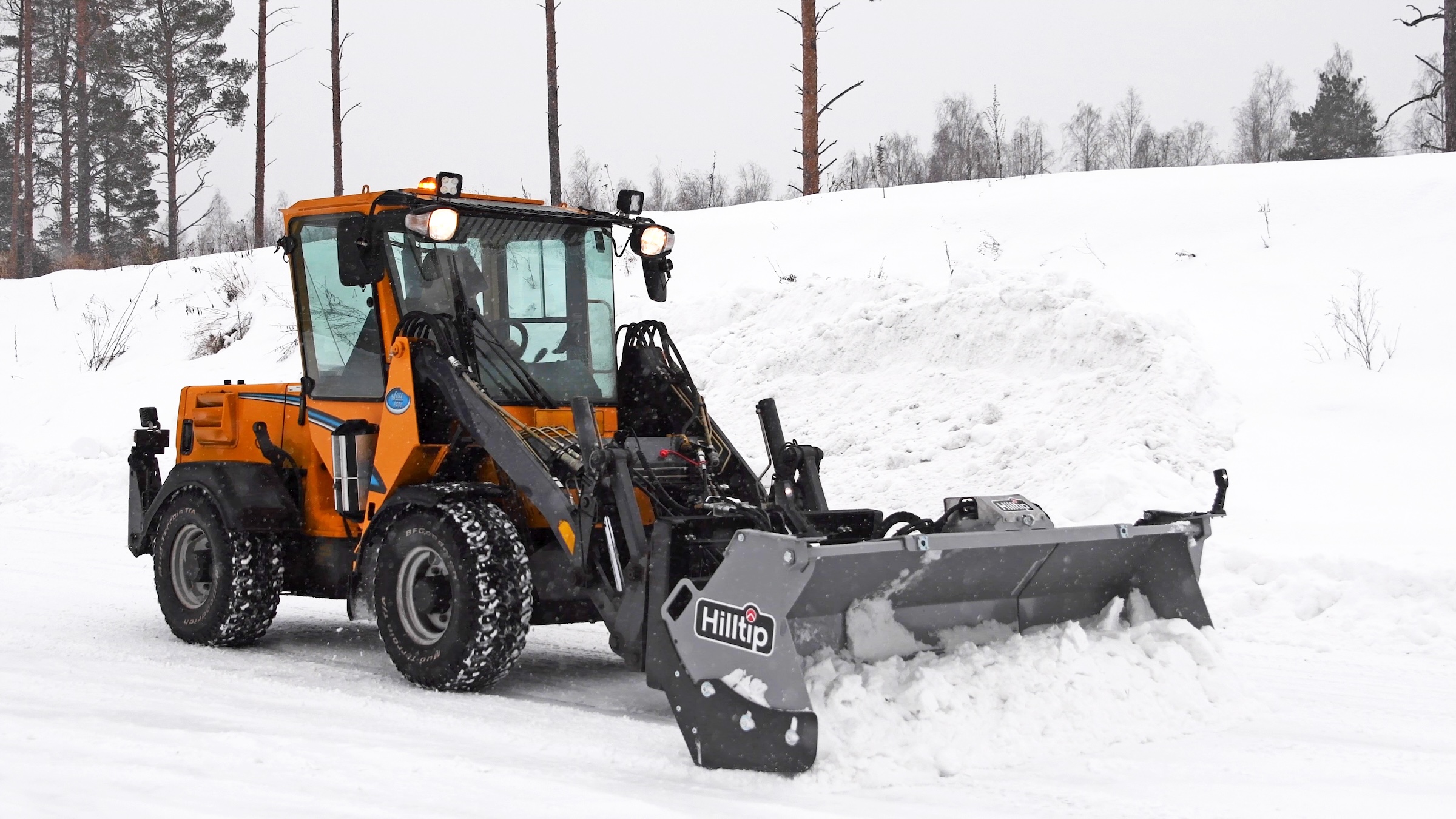 Orange wheel loader with Hilltip snow plow attachment clearing snow from a winter road
