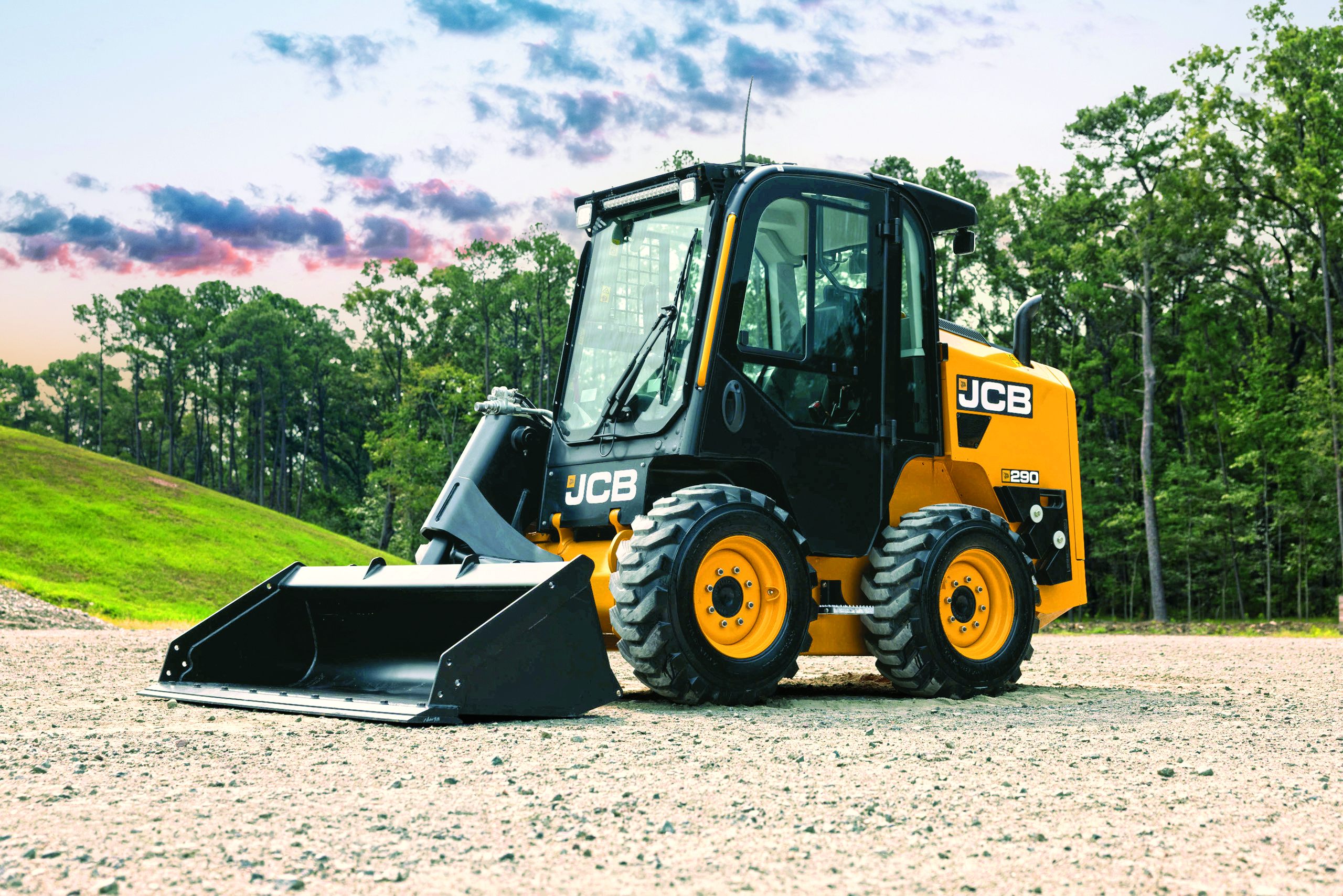 JCB skid steer loader with bucket attachment on gravel surface with trees and cloudy sky