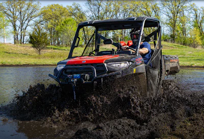 Red and black Polaris UTV splashing through muddy water with passengers wearing safety gear