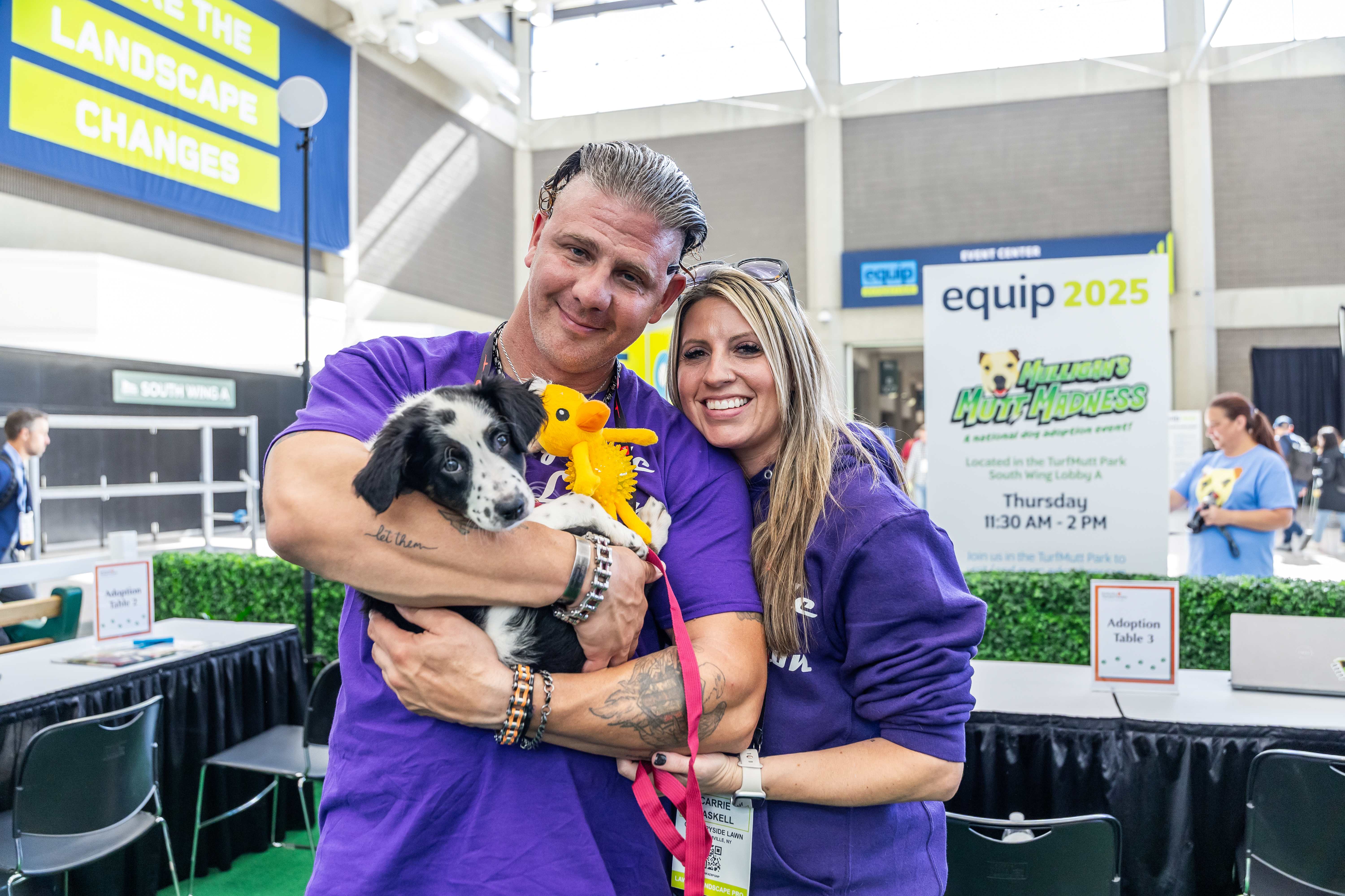 Two people in purple shirts holding a black and white dog at an equip 2025 event