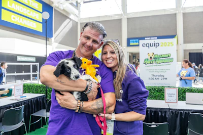 Two people in purple shirts holding a black and white dog at an equip 2025 event