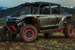 Custom gray and red off-road truck with elevated suspension parked in mountainous terrain with evergreen forest backdrop