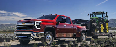 Red 2027 Chevy Silverado 3500 HD truck towing green John Deere tractor in rural farm setting
