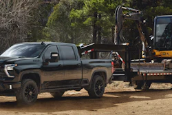 Black 2027 Chevrolet Silverado HD pickup truck towing a flatbed trailer with yellow construction excavator in a wooded area.