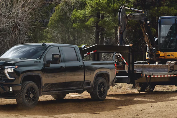 Black 2027 Chevrolet Silverado HD pickup truck towing a flatbed trailer with yellow construction excavator in a wooded area.