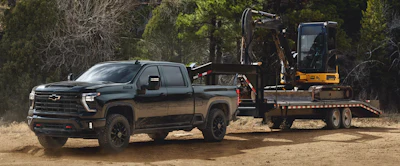 Black 2027 Chevrolet Silverado HD pickup truck towing a flatbed trailer with yellow construction excavator in a wooded area.
