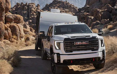 White 2027 GMC Sierra HD AT4 truck towing a white trailer through a rocky desert canyon landscape