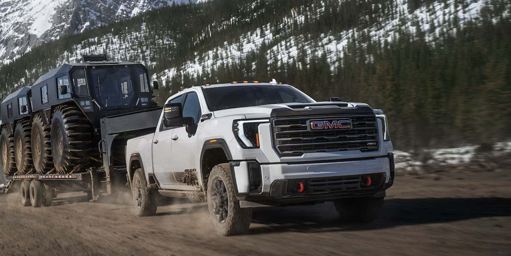 White 2027 GMC Sierra HD pickup truck towing large military trailer through snowy mountain terrain with forested slopes