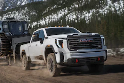 White 2027 GMC Sierra HD pickup truck towing large military trailer through snowy mountain terrain with forested slopes