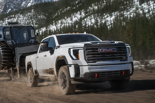 White 2027 GMC Sierra HD pickup truck towing large military trailer through snowy mountain terrain with forested slopes