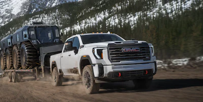 White 2027 GMC Sierra HD pickup truck towing large military trailer through snowy mountain terrain with forested slopes