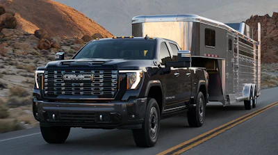 Black GMC Sierra truck towing a silver horse trailer on a desert highway with rocky mountains in background