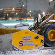 Yellow snow plow with Arctic Sectional Snow-Pusher attachment clearing snow from urban street at night